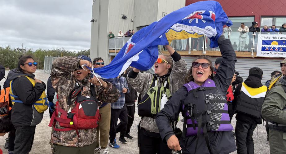 Trois jeunes souriants tiennent un drapeau tłı̨chǫ au-dessus de leurs têtes.