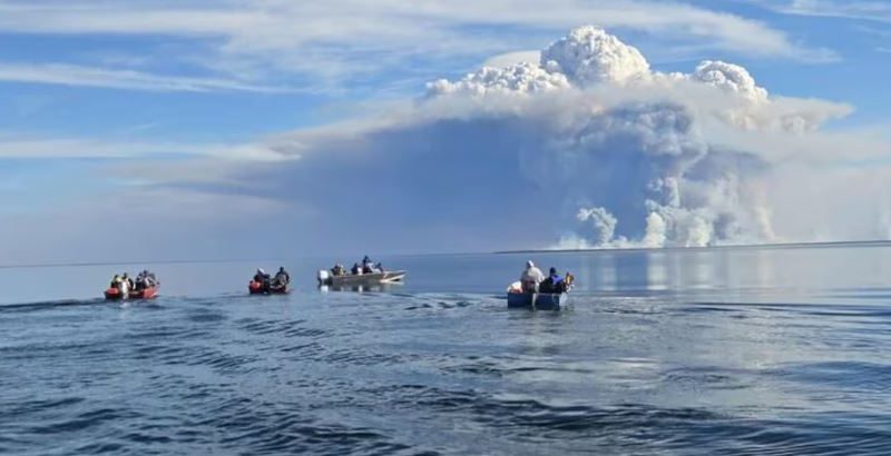 Des gens dans des embarcations sur un lac avec une colonne de fumée en arrière-plan.