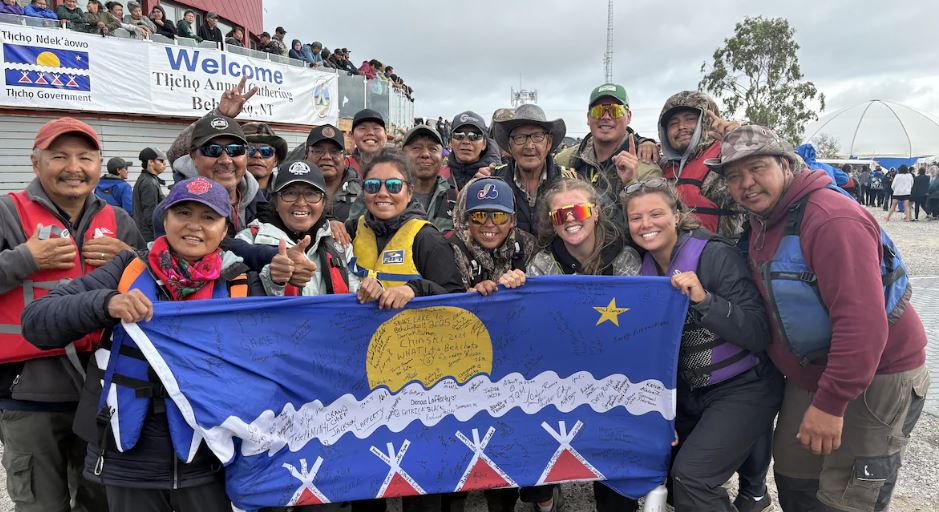 Une vingtaine de participants sourient vers l’appareil photo en tenant un drapeau tłı̨chǫ.