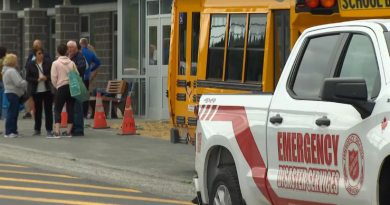 Un véhicule des services d’urgence est stationné devant une école où des personnes se rassemblent, tandis qu’un autobus scolaire et des cônes de sécurité indiquent une possible intervention.