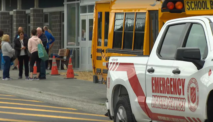 Un véhicule des services d’urgence est stationné devant une école où des personnes se rassemblent, tandis qu’un autobus scolaire et des cônes de sécurité indiquent une possible intervention.