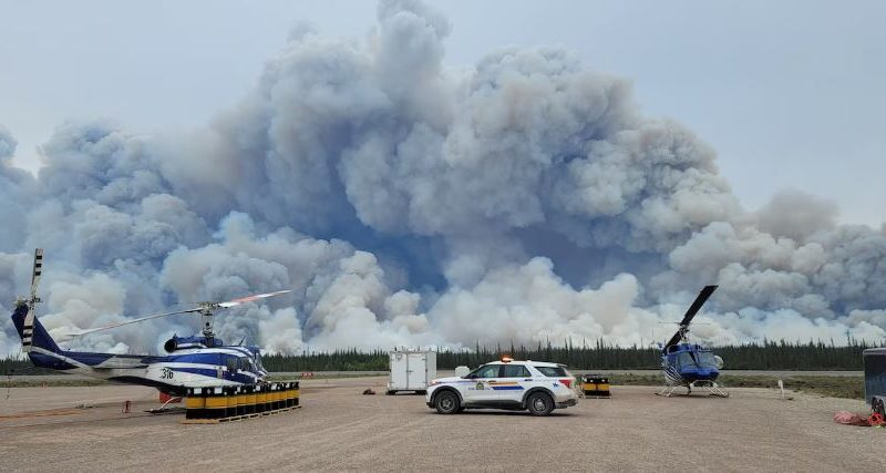 Une voiture de la GRC et deux hélicoptères sur un large terrain, alors qu'à l'horizon une épaisse fumée s'élève.