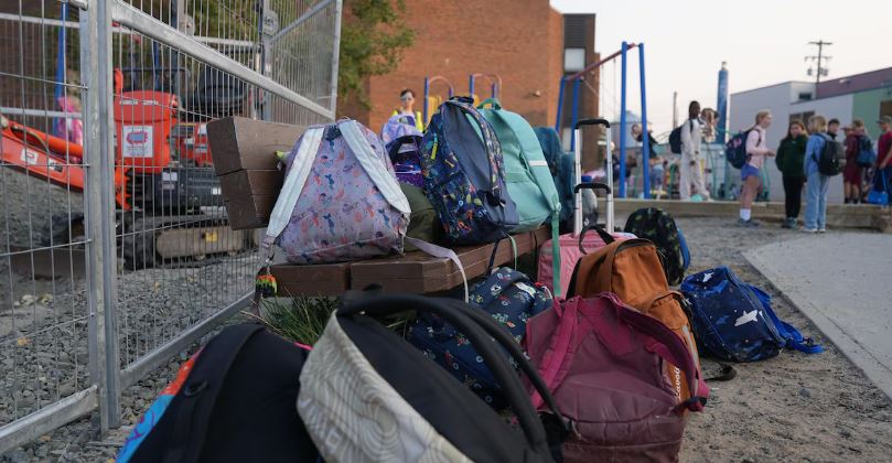 Des sacs à dos d'enfants entassés dans une cour d'école achalandée.