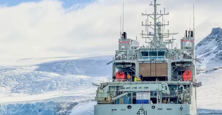 Un navire de la Marine canadienne est sur l'eau, sur une côte escarpée et glacée en Antarctique. On voit des gens sur le pont du navire.