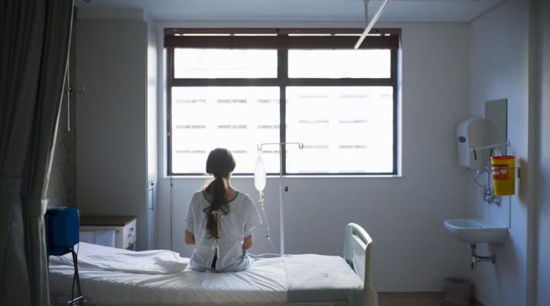 Une patiente de dos, assise sur un lit d'hôpital, regarde par la fenêtre.