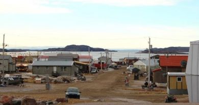 Une rue en terre à Kugaaruk bordée de bâtiments peu élevés et de plusieurs voitures et véhicules tout-terrain.