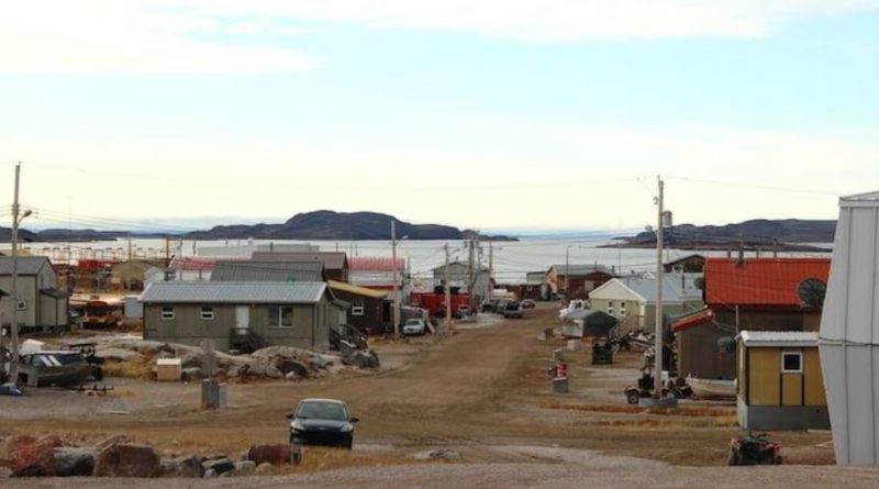 Une rue en terre à Kugaaruk bordée de bâtiments peu élevés et de plusieurs voitures et véhicules tout-terrain.