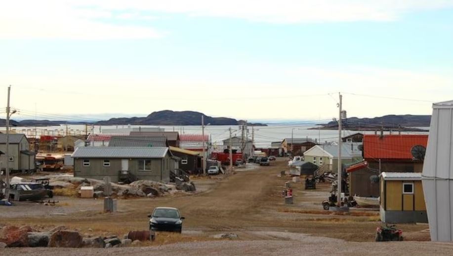 Une rue en terre à Kugaaruk bordée de bâtiments peu élevés et de plusieurs voitures et véhicules tout-terrain.