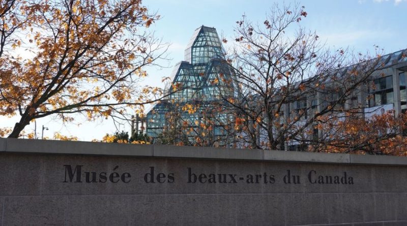 Le nom du musée est écrit sur un muret de briques. En arrière-plan, l'édifice du musée et des arbres avec quelques feuilles oranges.