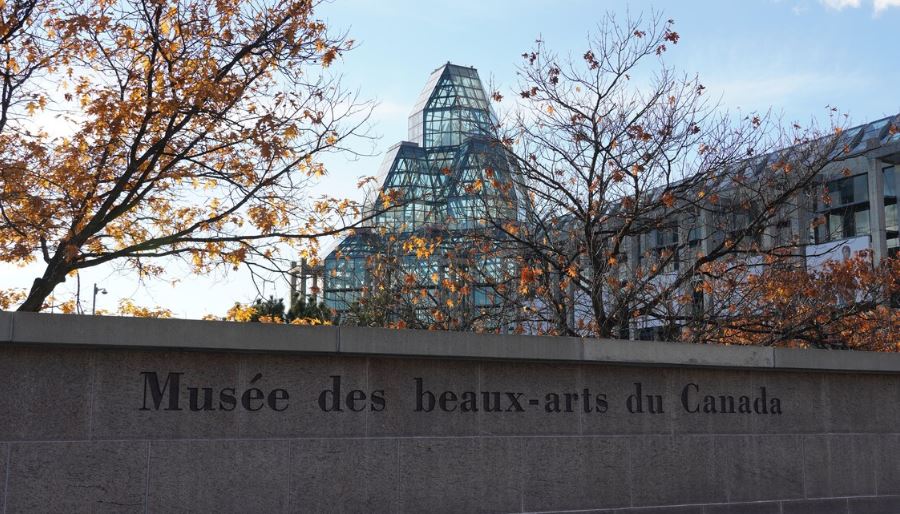 Le nom du musée est écrit sur un muret de briques. En arrière-plan, l'édifice du musée et des arbres avec quelques feuilles oranges.