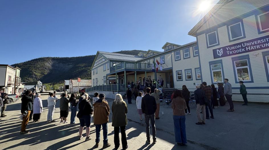 Des personnes dans la rue devant un bâtiment de l'Université du Yukon à Dawson regardent des personnes en train de signer des papiers.
