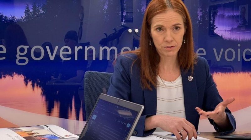 Une femme parle assise à une table couverte de piles de papiers et d'exemplaires du budget.